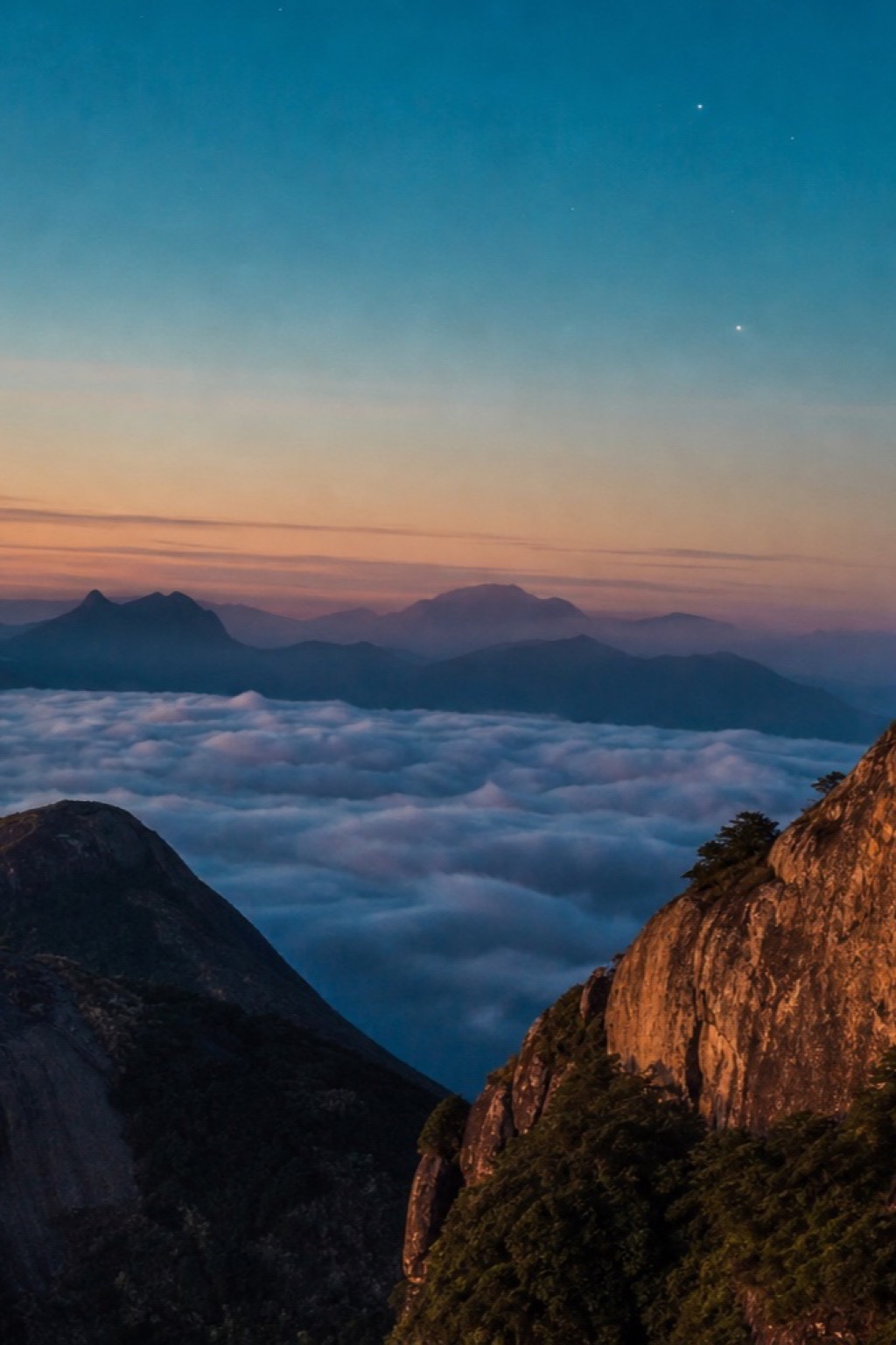 A agulha de granito do Dedo de Deus iluminada pelo poente sobre um mar de nuvens, contra um céu azul profundo