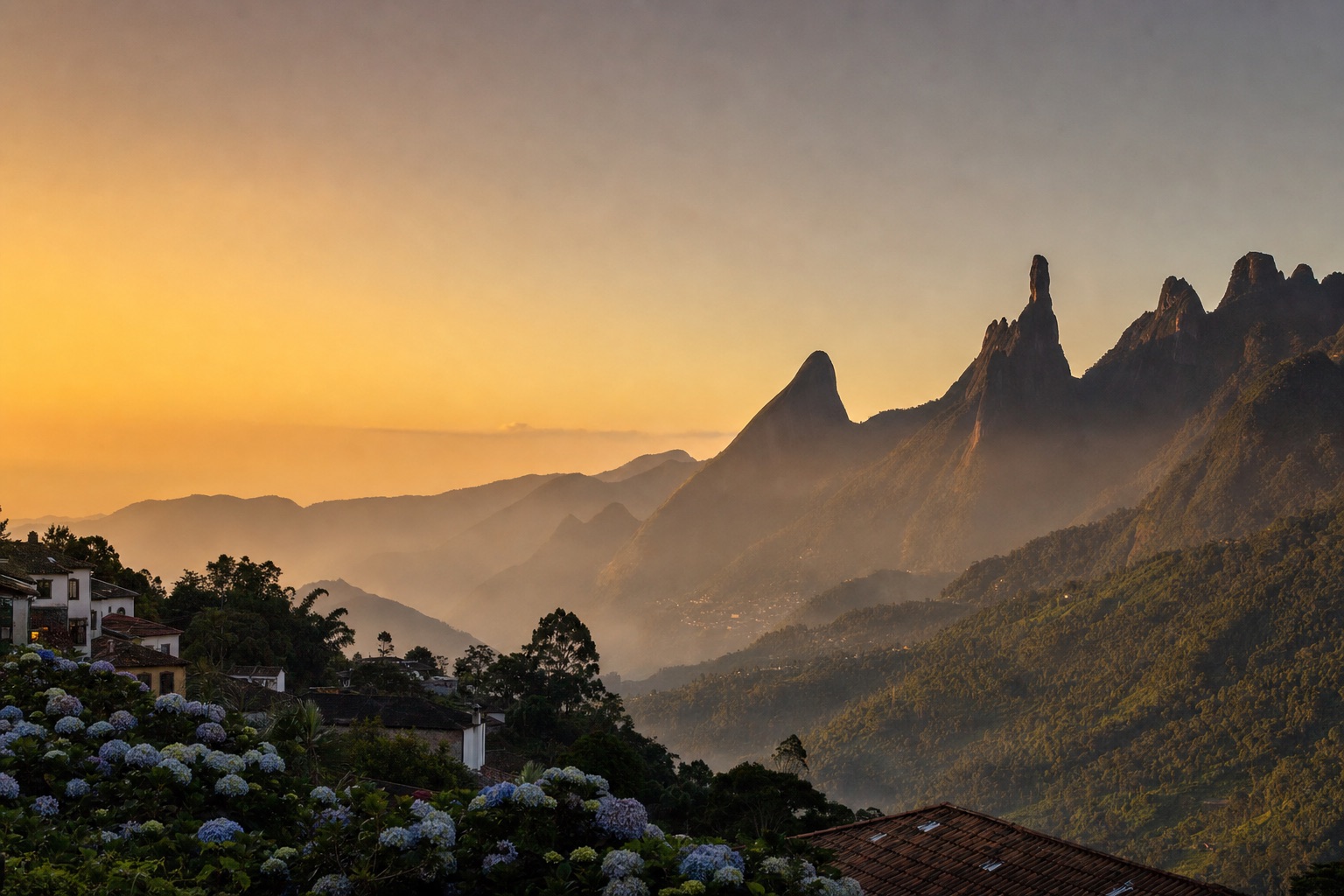 Panorama de la sierra al atardecer: los picos de granito de la Serra dos Órgãos sobre los valles llenos de niebla, con los tejados de la ciudad serrana en primer plano