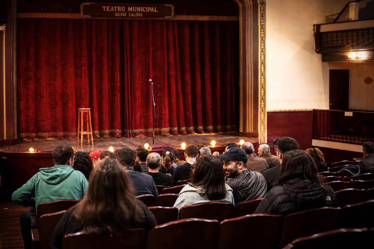 Sala de teatro histórica con butacas de madera, telón de terciopelo y público llegando antes de un espectáculo